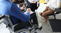 Disabled man with documents in hands sitting in wheelchair against background of colleagues closeup. Adaptation of disabled people and success to work concept